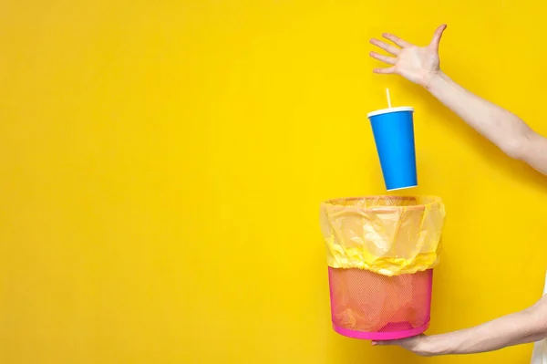 hand throws plastic cup into trash can on yellow background, ecology concept, garbage falls into trash can, copy space