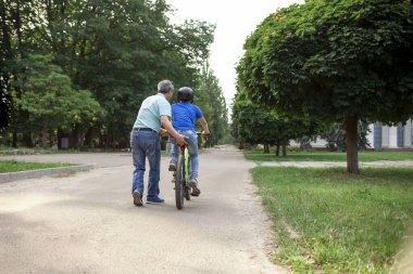 Asyalı baba oğluna bisiklet sürmeyi öğretiyor, Koreli son sınıf öğrencisi çocuğa yardım ediyor, kasklı çocuk yazın babasıyla birlikte parkta dinleniyor.
