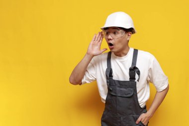Üniformalı Asyalı erkek müteahhit, sarı izole edilmiş arka plan hakkında bilgi veriyor. Koreli ustabaşı sert şapka takıp bağırıyor ve konuşuyor.