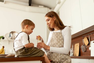 Önlüklü genç anne, evdeki mutfakta yemesi için küçük oğluna kurabiye ve süt veriyor, 2 yaşındaki çocuk ailesiyle kahvaltı ediyor, kadın çocuğu besliyor.