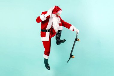 Güneş gözlüklü ve takım elbiseli Noel Baba mavi arka planda kaykayla zıplar ve uçar. Hipster Noel Baba kaykaya biner ve yılbaşı ve yılbaşı için havada numara yapar.