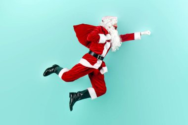 Bir çanta dolusu hediyeyle Noel Baba atlıyor ve mavi izole edilmiş arka planda hızla havada uçuyor. Noel Baba uçuyor ve yeni yıl için acele ediyor.