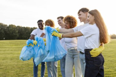 Çöp torbalarıyla eldiven giyen çok ırklı bir grup gönüllü parkta çöp ve plastik topluyor, öğrencilerden oluşan bir grup gezegene yardım ediyor, gençlik çevreyle ilgileniyor.