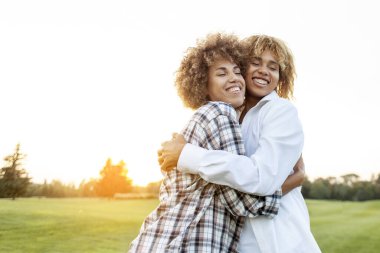 Günbatımında parkta gülümseyen ve sarılan iki Afro-Amerikan kıvırcık kadın, kız kardeşler ve arkadaşları sevgi ve ilgi gösteriyorlar.