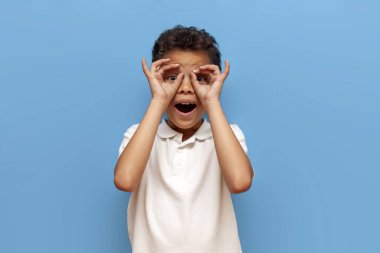 Afro-Amerikalı genç çocuk elleriyle dürbünle bakar ve mavi izole edilmiş arka planda şaşırır, çocuk elleriyle gözlük ve maske tasvir eder ve hayretle bakar.