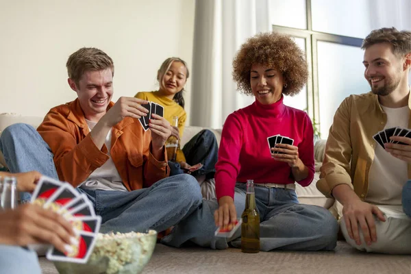 multiracial group of young people sitting at home with beer and popcorn and playing cards with friends, students of different ethnicities playing gambling board games at a party and having fun