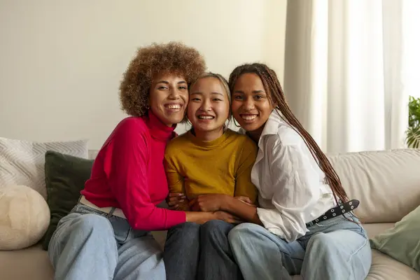African American women and Asian woman sitting on sofa at home and hugging, interracial girlfriends smiling and looking at camera