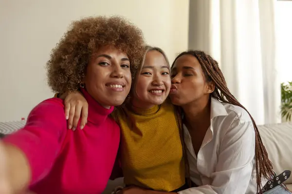 African American women and Asian woman sitting on sofa at home taking selfie and hugging, interracial girlfriends smiling and taking photos on phone