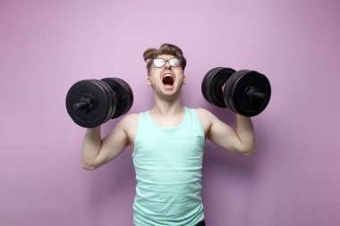 young confident guy student lifts heavy dumbbells on a pink background, motivated nerd in glasses goes in for sports and shouts