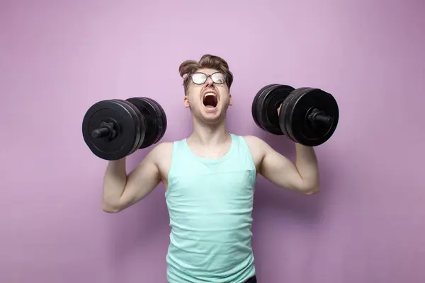 stock image young confident guy student lifts heavy dumbbells on a pink background, motivated nerd in glasses goes in for sports and shouts