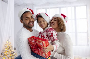 Noel Baba şapkalı Afro-Amerikan aile Noel ağacının arka planında sarılıp gülümsüyor, genç çocuk hediye kutusunu tutuyor ve kış tatilini ailesiyle birlikte kutluyor.