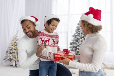 Noel Baba şapkalı Afro-Amerikan aile Noel ağacının arka planında sarılıp gülümsüyor, genç çocuk hediye kutusunu tutuyor ve kış tatilini ailesiyle birlikte kutluyor.