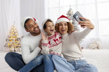 Afro-Amerikan ailesi Noel 'i çocuğuyla kutluyor ve akıllı telefonlarıyla selfie çekiyor, Noel Baba şapkalı çocuk internette fotoğraf çekiyor ve selamlaşırken elini sallıyor.