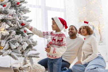 Afro-Amerikan ailesi Noel 'i çocuklarıyla kutluyor ve Noel ağacını evde süslüyor, Noel Baba şapkalı 10 yaşındaki çocuk dekoratif oyuncaklar tutuyor ve