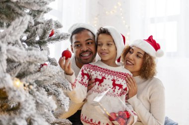 Afro-Amerikan ailesi Noel 'i çocuklarıyla kutluyor ve Noel ağacını evde süslüyor, Noel Baba şapkalı 10 yaşındaki çocuk dekoratif oyuncaklar tutuyor ve