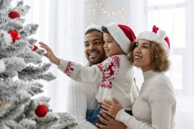 Afro-Amerikan ailesi Noel 'i çocuklarıyla kutluyor ve Noel ağacını evde süslüyor, Noel Baba şapkalı 10 yaşındaki çocuk dekoratif oyuncaklar tutuyor ve