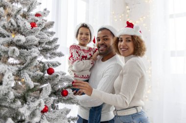 Afro-Amerikan ailesi Noel 'i çocuklarıyla kutluyor ve Noel ağacını evde süslüyor, Noel Baba şapkalı 10 yaşındaki çocuk dekoratif oyuncaklar tutuyor ve
