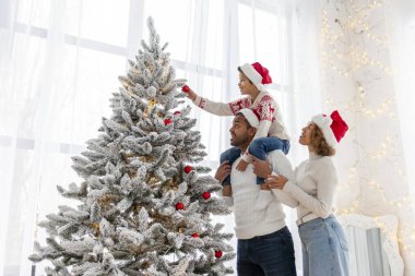 Afro-Amerikan ailesi Noel 'i çocuklarıyla kutluyor ve Noel ağacını evde süslüyor, Noel Baba şapkalı 10 yaşındaki çocuk dekoratif oyuncaklar tutuyor ve