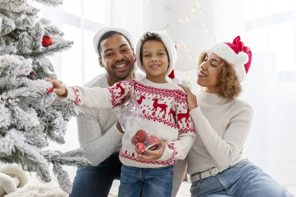 African-American family celebrates Christmas with their child and decorates the Christmas tree at home, 10-year-old boy with his parents in Santa hat holds decorative toy and helps