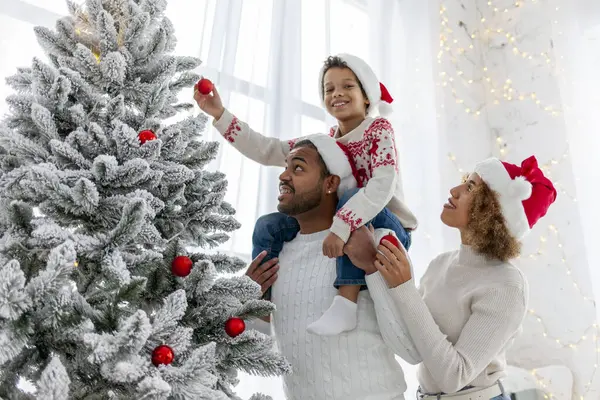 Afro-Amerikan ailesi Noel 'i çocuklarıyla kutluyor ve Noel ağacını evde süslüyor, Noel Baba şapkalı 10 yaşındaki çocuk dekoratif oyuncaklar tutuyor ve