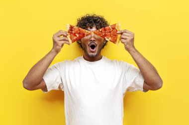 Aç Hintli adam elinde iki dilim lezzetli pizza tutuyor ve sarı izole edilmiş arka planda şaşırıyor. Şaşırmış Hindu adam yiyor ve fast food reklamı yapıyor.