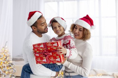Noel Baba şapkalı Afro-Amerikan aile Noel ağacının arka planında sarılıp gülümsüyor, genç çocuk hediye kutusunu tutuyor ve kış tatilini ailesiyle birlikte kutluyor.