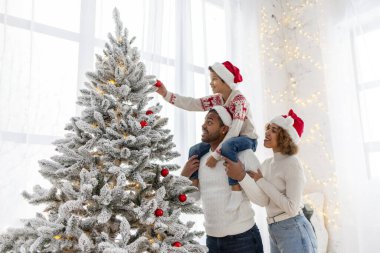 Afro-Amerikan ailesi Noel 'i çocuklarıyla kutluyor ve Noel ağacını evde süslüyor, Noel Baba şapkalı 10 yaşındaki çocuk dekoratif oyuncaklar tutuyor ve