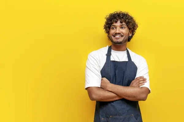 young guy Indian waiter in apron smiling crossing his arms on yellow isolated background, male Hindu barista in uniform looking at copy space