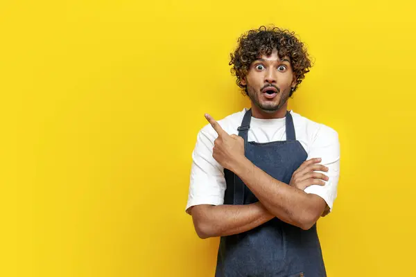 young surprised guy indian waiter in apron pointing at copy space on yellow isolated background, male indian barista in uniform showing and advertising in amazement