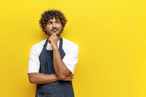 young thoughtful guy Indian waiter in apron plans and thinks on yellow isolated background, puzzled male Indian barista in uniform imagines and dreams