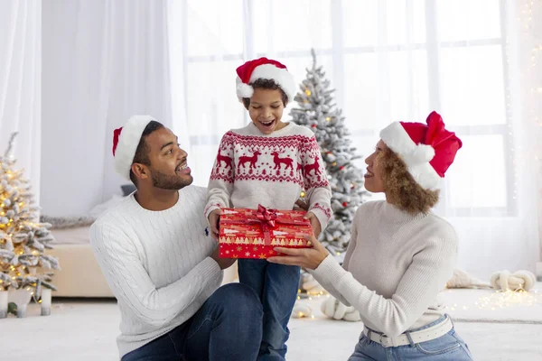 African-American family in Santa hats hugging and smiling at home against the background of christmas tree, teenage boy holds gift box and celebrates the winter holidays with his parents together