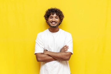 Sarı izole arka planda çapraz kolları olan genç Hintli kıvırcık adam ve gülümseyen beyaz tişörtlü Güney Asyalı adam kameraya bakıyor.