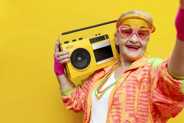 funny old granny with tape recorder in sports hipster clothes takes selfie online on yellow isolated background, elderly woman in stylish youth clothes listens to loud music and dances at disco