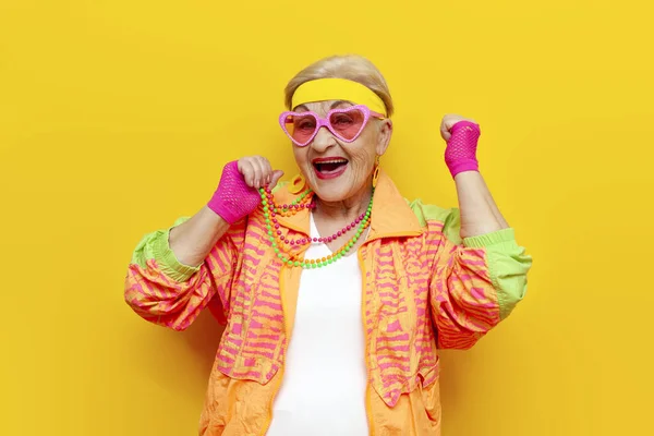 funny crazy old granny in sports hipster clothes rejoices at success and victory on a yellow isolated background, elderly woman in youth clothes celebrates success and wins