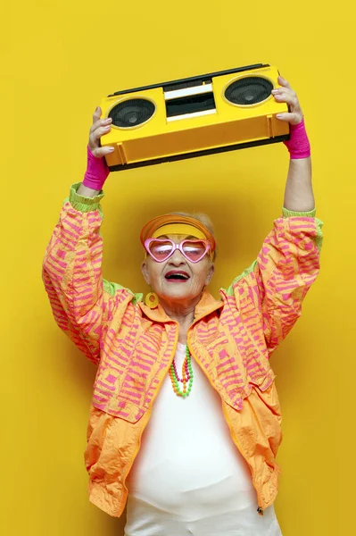 funny crazy old granny with tape recorder in sports hipster clothes listens to music and sings on yellow isolated background, elderly woman in stylish youth clothes with record player screams at party
