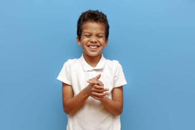 Beyaz polo giyen Afrikalı Amerikalı çocuk mavi arka planda gülüyor ve alkışlıyor, 10 yaşındaki çocuk gülümsüyor ve ellerini çırpıyor.