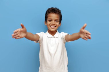 Beyaz polo giyen Afro-Amerikalı çocuk kucaklaşmak istiyor ve ellerini mavi izole arka planda ileri çekiyor, 10 yaşındaki çocuk önünde boş elleriyle duruyor.