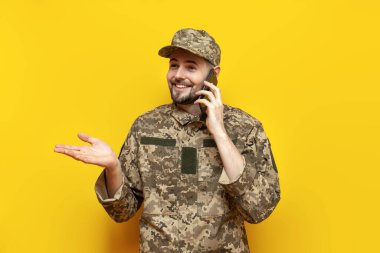 Piksel kamuflaj üniformalı genç Ukraynalı erkek asker sarı izole edilmiş bir arka planda telefonla konuşuyor. Ukrayna ordusunun askeri öğrencisi cep telefonuyla iletişim kuruyor.