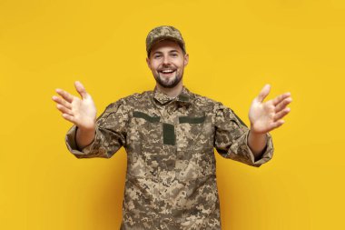 Kamuflaj üniformalı genç Ukraynalı asker sarı izole bir arka planda kucaklaşmayı ve hoş geldin demeyi gösteriyor. Ukrayna ordusunun askeri öğrencisi önünde boş elleriyle duruyor.