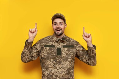 Kamuflaj piksel üniformalı genç Ukraynalı erkek asker ellerini sarı izole bir arka plana doğrultuyor. Ukrayna ordusunun askeri öğrencisi yukarıdan kopya uzay reklamları yapıyor.
