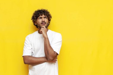 Beyaz tişörtlü genç dalgın Hintli adam sarı yalıtılmış arka plan üzerinde düşünüyor ve hayal kurup hayal kuruyor. Yüksek kalite fotoğraf