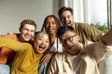 Kameraya gülümseyen ve selfie çeken çok ırklı genç arkadaşlar, Asyalı, Afrikalı, Amerikalı ve Avrupalı öğrenciler kucaklaşıp fotoğraf çekiyorlar.