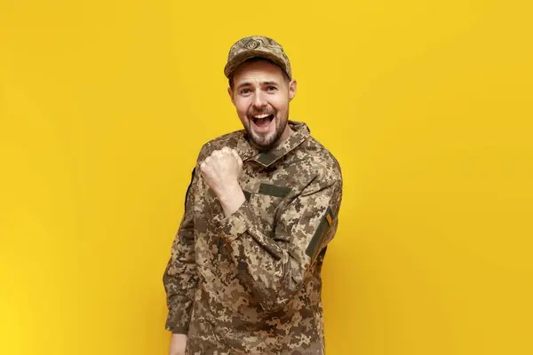 young Ukrainian male soldier in pixel camouflage uniform celebrates victory on a yellow isolated background, guy military cadet of the Ukrainian army wins and rejoices at luck and success