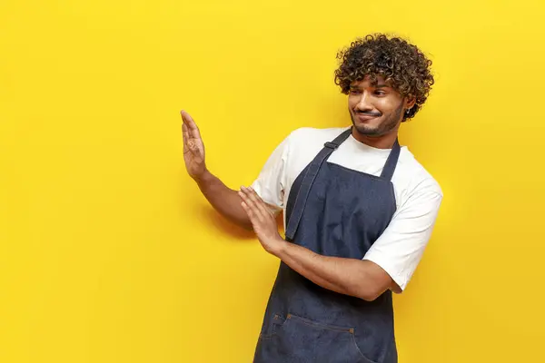 young indian male barista in an apron refuses and shows disgust on a yellow isolated background, guy indian waiter in uniform rejects and avoids