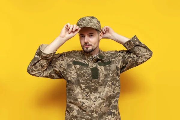 young Ukrainian male soldier in a pixel camouflage uniform puts on a cap on a yellow isolated background, the guy is a military cadet of the Ukrainian army