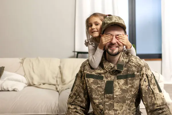 young dad, a soldier of the Ukrainian army in uniform, returned home to his family and child, a little girl daughter closes her eyes to her military father and greets him with a surprise