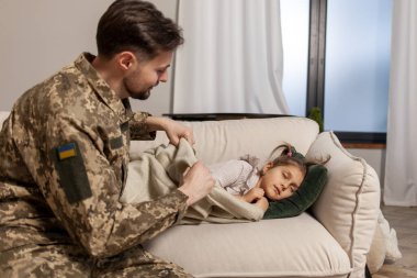Ukrayna ordusunun genç askeri kamuflaj üniforması içinde evine, çocuğuna döndü. Bir asker babası küçük kızını evinde kanepede örterek öptü. Askeri Ukrayna konsepti.