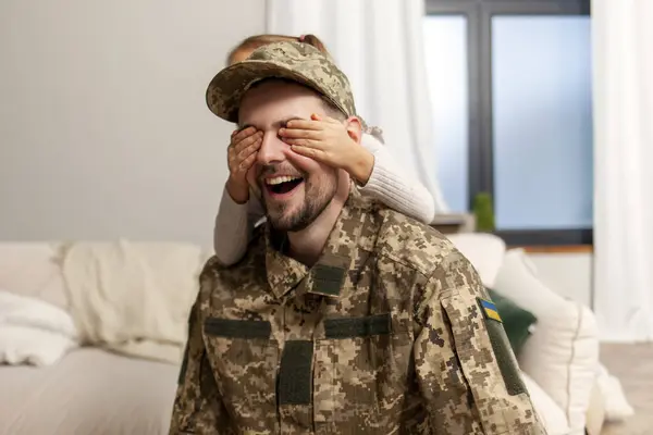 young dad, a soldier of the Ukrainian army in uniform, returned home to his family and child, a little girl daughter closes her eyes to her military father and greets him with a surprise