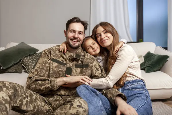 young soldier of the Ukrainian army in a camouflage uniform returned home to his family, a military dad hugs his little daughter and wife at home, a happy military family sits at home on the sofa and