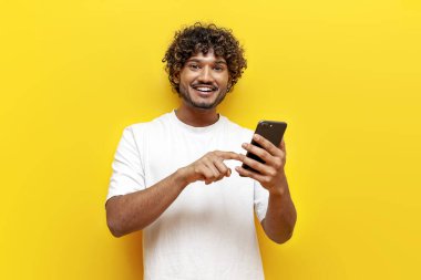 neşeli Hintli adam akıllı telefon kullanıyor ve sarı izole edilmiş arka planda interneti seçiyor, kıvırcık adam telefonda mesaj yazıyor ve bağlanıyor